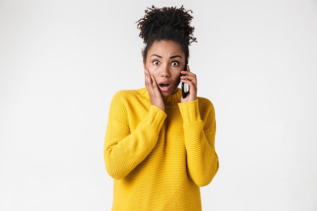 Imagem de uma bela jovem africana animado emocional chocado mulher posando sobre uma parede branca, falando por telefone celular.