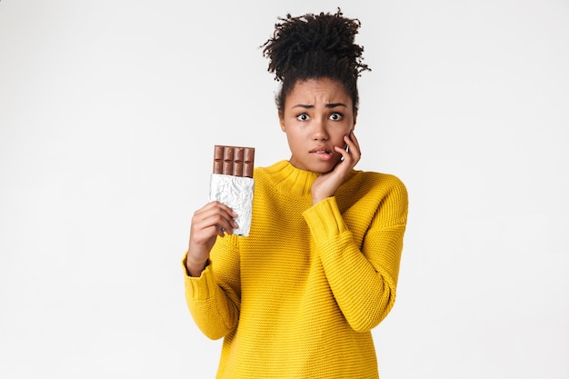 Foto imagem de uma bela jovem africana confusa mulher posando sobre uma parede branca segurando doces de chocolate.