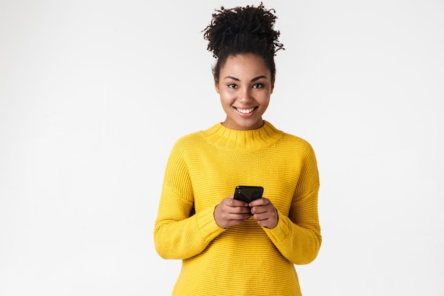 Foto imagem de uma bela jovem africana feliz posando sobre uma parede branca, usando telefone celular.