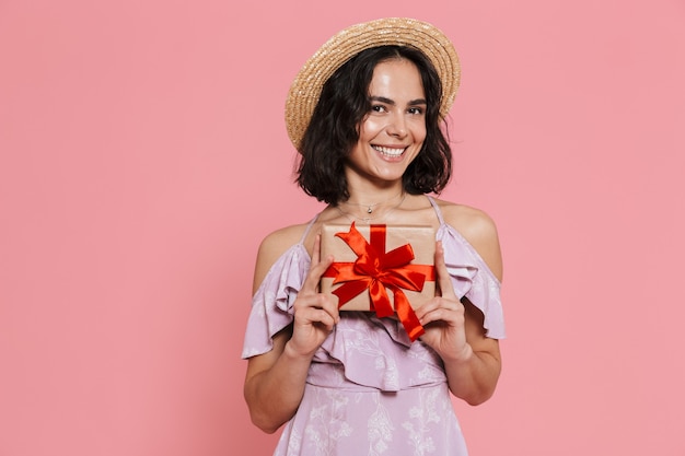 Imagem de uma bela jovem feliz posando isolada sobre uma parede rosa segurando uma caixa de presente.