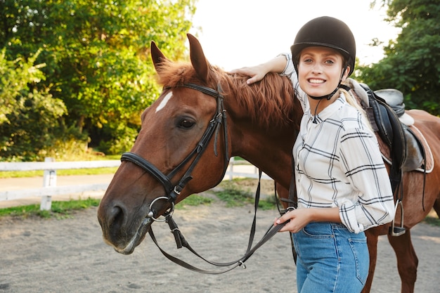 Imagem de uma bela jovem loira com chapéu parado ao lado do cavalo no quintal no campo