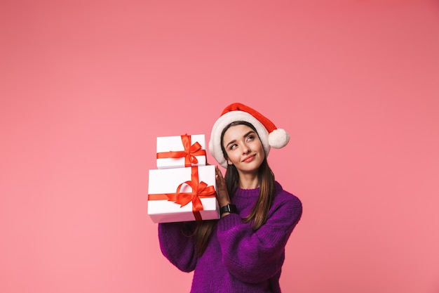 Imagem de uma bela jovem pensando posando isolado sobre rosa segurando caixas de presente com chapéu de Natal.