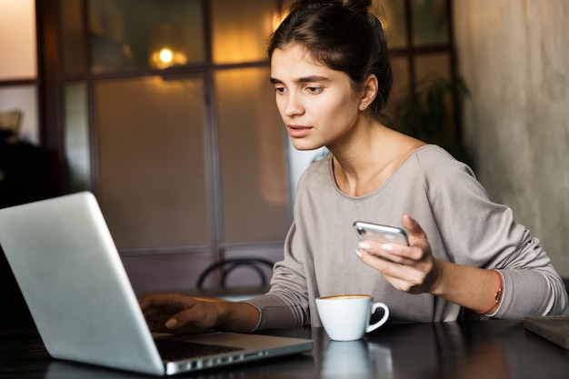 Imagem de uma bela jovem sentada em um café dentro de casa, usando o computador portátil e o telefone celular.