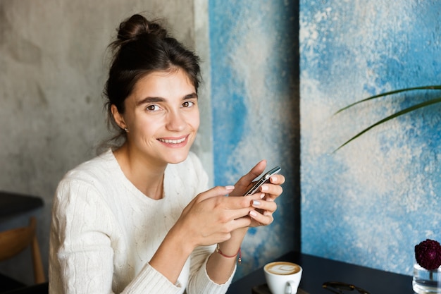 Imagem de uma bela jovem sentada no café, bebendo café dentro de casa, usando o telefone móvel conversando.