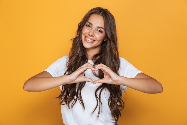 Foto imagem de uma charmosa mulher de 20 anos com cabelo comprido sorrindo e mostrando formato de coração com os dedos, isolada sobre parede amarela