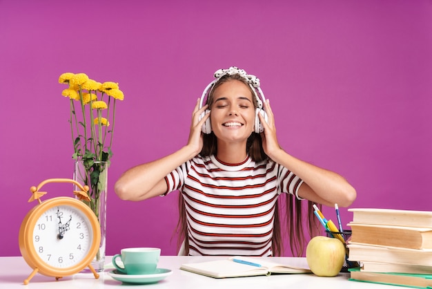 Imagem de uma garota feliz ouvindo música com fones de ouvido e sorrindo enquanto está sentado na mesa, isolado sobre a parede violeta