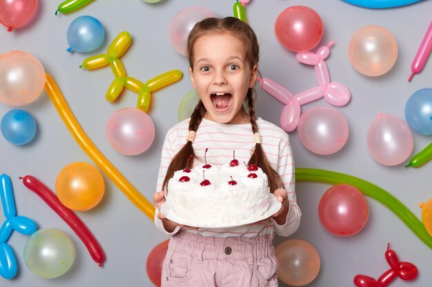 Imagem de uma garotinha extremamente feliz espantada com tranças vestindo roupas casuais posando isolada sobre fundo cinza com balões segurando bolo de aniversário