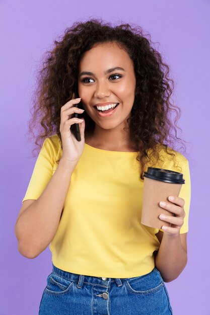 Imagem de uma jovem afro-americana com cabelo encaracolado, segurando uma xícara de café e falando em um smartphone, isolada sobre a parede violeta