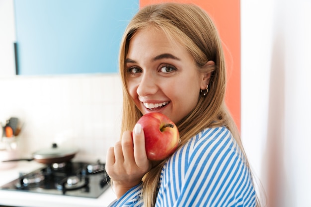 Imagem de uma jovem alegre vestindo uma camisa listrada, sorrindo e comendo maçã na cozinha
