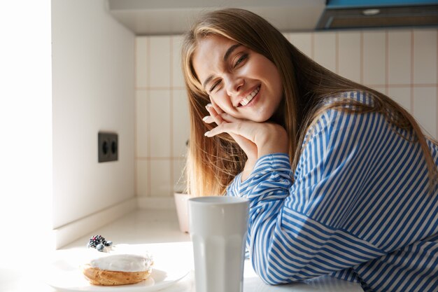 Foto imagem de uma jovem atraente vestindo camisa listrada sorrindo e bebendo chá na cozinha