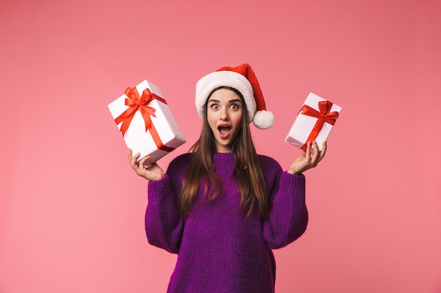 Imagem de uma jovem chocada emocional posar isolado sobre o espaço rosa segurando caixas de presente com chapéu de natal.