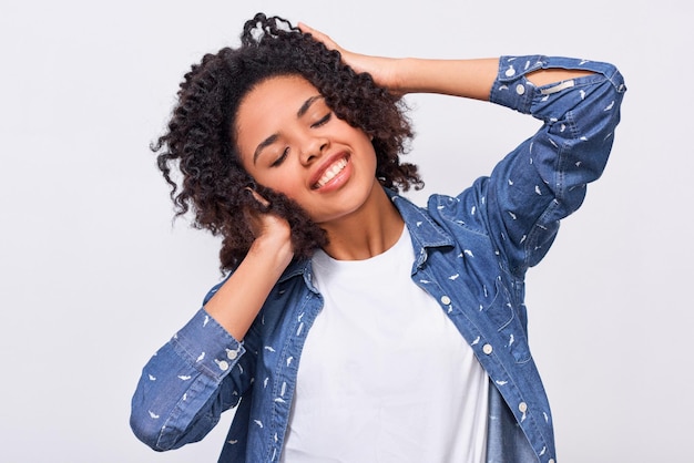 Imagem de uma jovem de pele escura satisfeita, vestida de camisa azul, segurando as mãos na cabeça, sente-se feliz mulher afro-americana sorrindo amplamente e sentindo-se satisfeita posando sobre parede branca