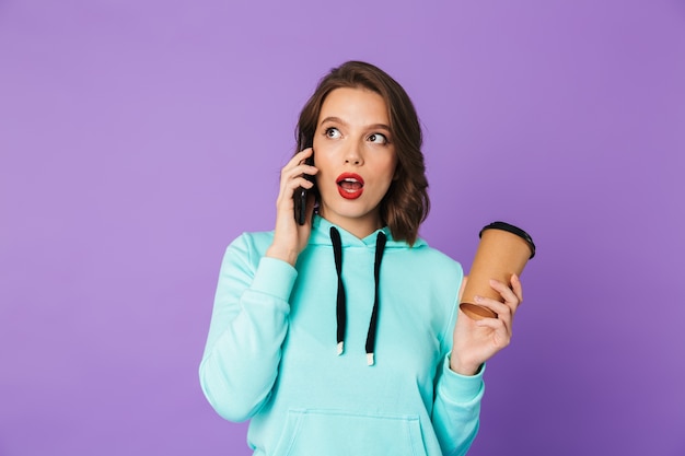 Imagem de uma jovem emocional chocada posando isolada sobre a parede roxa, falando por telefone celular.