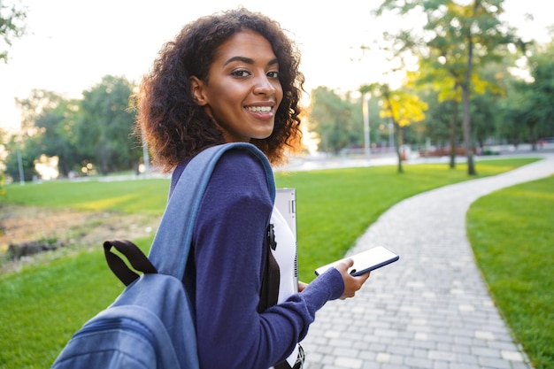 Imagem de uma jovem estudante africana bonita andando no parque conversando por telefone celular.