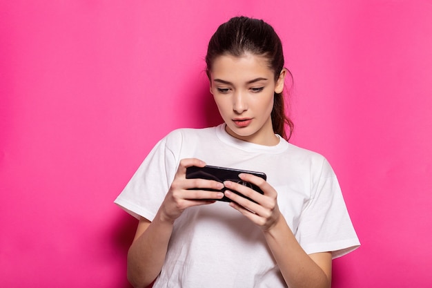 Imagem de uma jovem feliz em uma camiseta branca, jogando no celular e ganhando, regozijando-se em pé sobre um fundo rosa