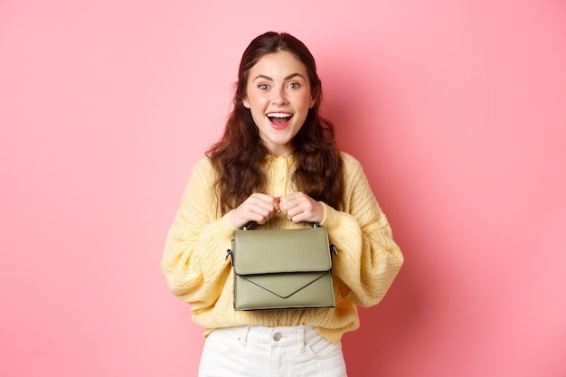 Imagem de uma jovem feliz indo às compras, segurando a bolsa e sorrindo animada, pronta para ir, encostada na parede rosa