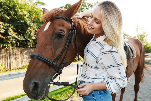 Imagem de uma jovem loira alegre sorrindo e parada a cavalo no quintal no campo