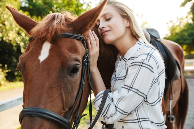 Imagem de uma jovem loira alegre sorrindo e parada a cavalo no quintal no campo