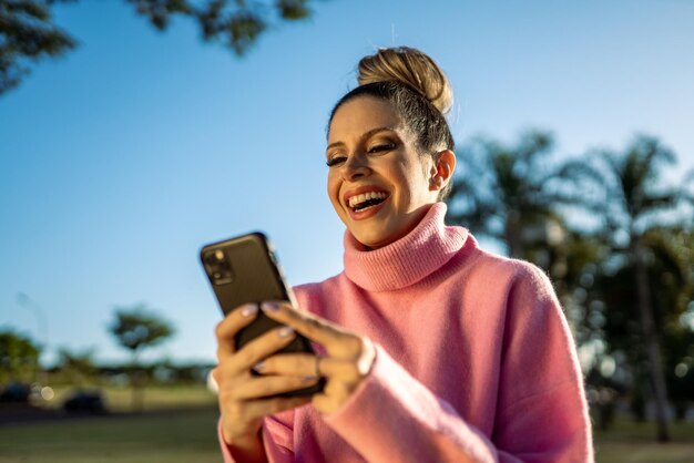 Imagem de uma jovem loira latina animada feliz usando o celular no fundo do parque