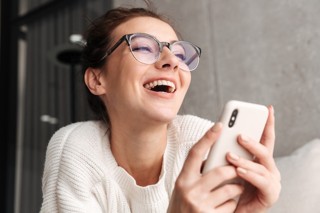 Foto imagem de uma jovem morena positiva de óculos sorrindo e segurando o celular em casa