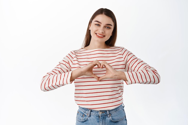 Imagem de uma jovem mulher bonita mostra coração, eu te amo, sinal, expressar sentimentos ternos sinceros, afeto, em pé sobre um fundo branco