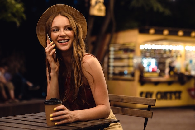 Imagem de uma jovem mulher feliz sentada no café ao ar livre à noite, bebendo café, falando por telefone celular.