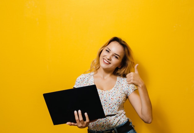 Imagem de uma jovem mulher segurando um laptop e feliz sobre parede amarela