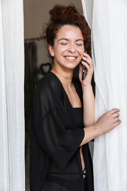 Imagem de uma jovem sorridente otimista em trajes de banho de lingerie posando dentro de casa em casa hotel perto de cortina falando por telefone celular.