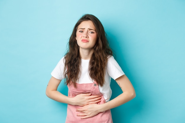 Imagem de uma jovem triste, carrancuda, de mãos dadas na barriga, tendo dores, cólicas menstruais dolorosas, em pé contra um fundo azul.