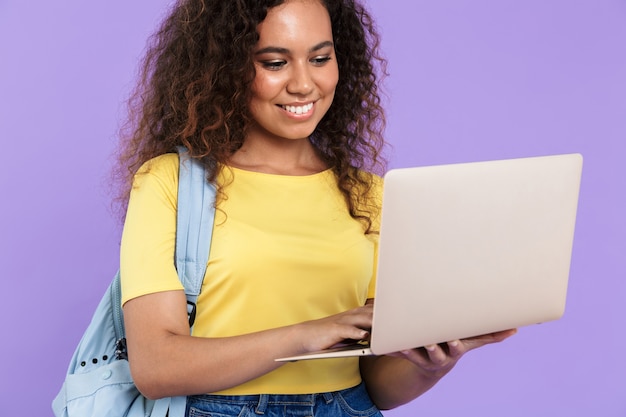 Foto imagem de uma linda aluna afro-americana com cabelo encaracolado segurando um laptop isolado sobre a parede violeta