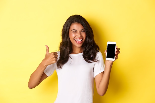 Imagem de uma linda garota afro-americana piscando e sorrindo, mostrando a tela do smartphone e o polegar para cima ...