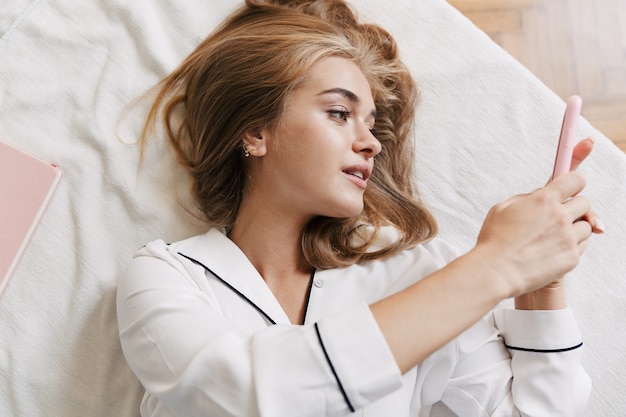 Imagem de uma linda jovem de pijama branco segurando o celular enquanto está deitada na cama em casa