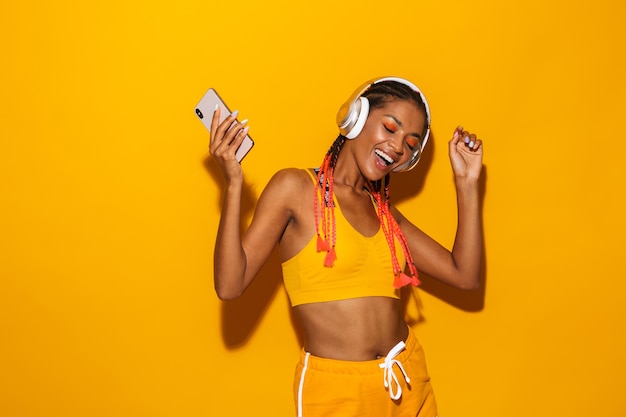 Imagem de uma linda mulher afro-americana cantando enquanto ouve música com smartphone e fones de ouvido isolados sobre a parede amarela