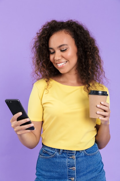 Imagem de uma linda mulher afro-americana com cabelo encaracolado segurando um smartphone e uma xícara de café isolada sobre a parede violeta