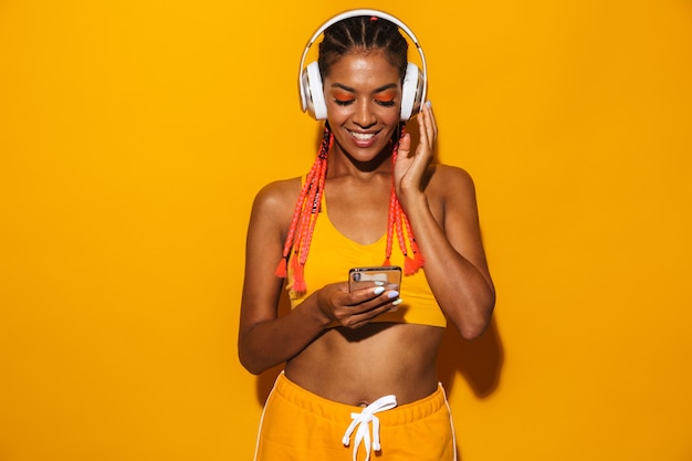 Imagem de uma linda mulher afro-americana usando tranças afro usando smartphone e fones de ouvido isolados sobre a parede amarela