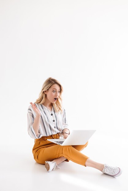 Imagem de uma linda mulher animada vestindo roupas casuais, olhando para o laptop enquanto está sentado no chão, isolado sobre uma parede branca