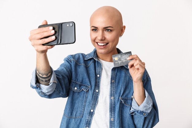 Imagem de uma linda mulher careca feliz posando isolado, falando pelo celular, tirar uma selfie segurando o cartão de crédito.