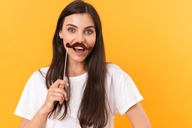 Imagem de uma linda mulher morena vestindo roupas básicas se divertindo e segurando um bigode de papel isolado sobre a parede amarela