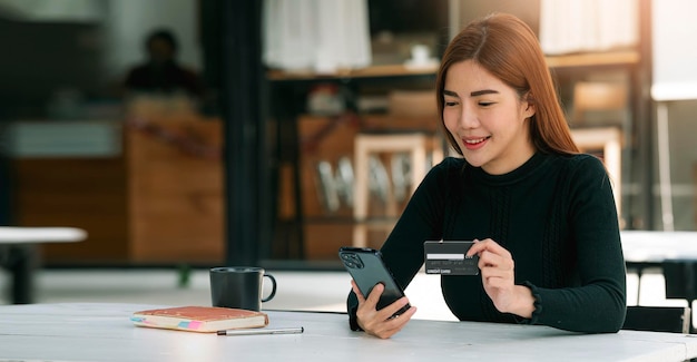 Imagem de uma linda mulher morena vestindo suéter preto segurando cartão de crédito e celular, fazendo compras online enquanto se senta em cafés,