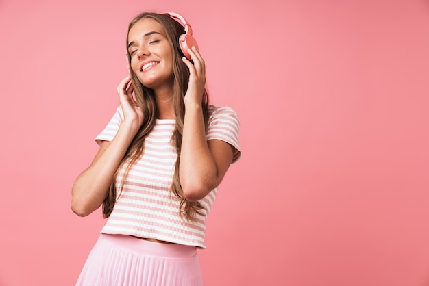 Imagem de uma linda mulher satisfeita vestindo roupas listradas, sorrindo e ouvindo música com fones de ouvido isolados sobre a parede rosa