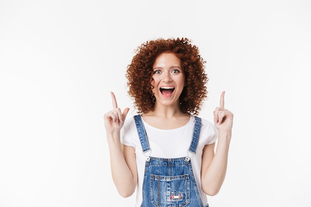 Imagem de uma linda ruiva encaracolada feliz posando isolado sobre a parede branca apontando.