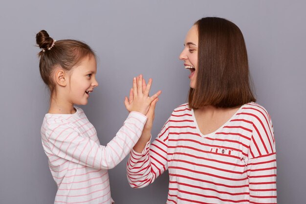 Imagem de uma mãe extremamente feliz e uma filha pequena vestindo camisas de estilo casual fazendo gestos de mais cinco tendo feito todas as coisas perfeitamente posando isoladas sobre fundo cinza