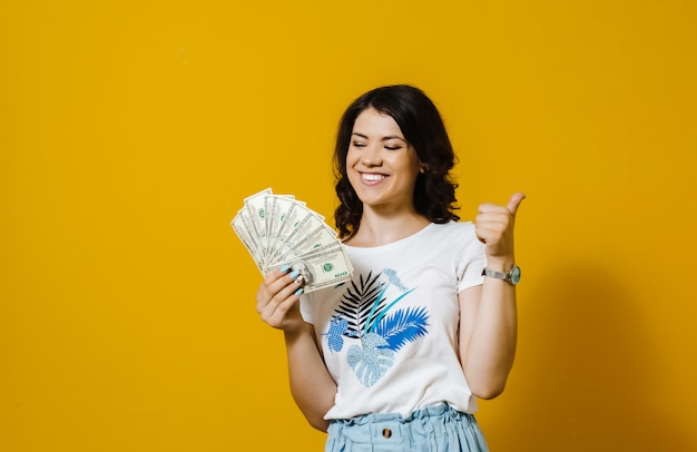 Imagem de uma menina sorridente segurando o monte de notas de dinheiro e mostrando okey gesto isolado sobre parede amarela