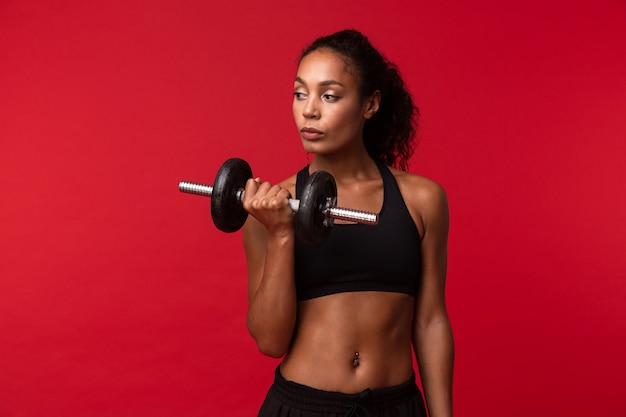 Imagem de uma mulher afro-americana de ginástica em roupa esportiva preta levantando halteres, isolada sobre uma parede vermelha