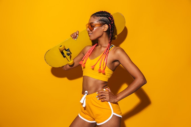 Imagem de uma mulher afro-americana feliz usando óculos escuros, segurando um skate e sorrindo, isolada sobre a parede amarela