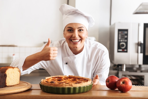 Imagem de uma mulher alegre, chef de uniforme branco, posando na cozinha em um café com assados