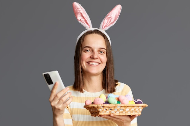 Imagem de uma mulher alegre e atraente usando orelhas de coelho segurando ovos de Páscoa coloridos e telefone celular olhando para a câmera usando um telefone inteligente isolado em fundo cinza