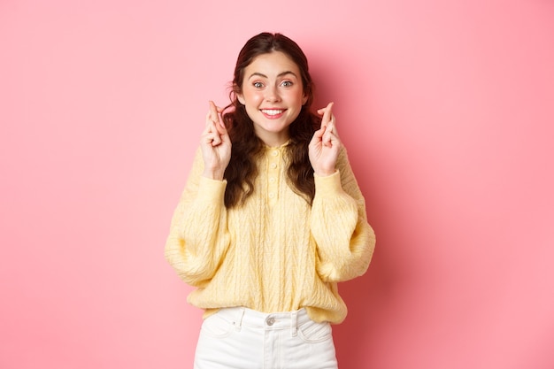 Imagem de uma mulher alegre e fofa cruzando os dedos para dar sorte e olhando esperançosa para a câmera fazendo um desejo em pé sobre a parede rosa