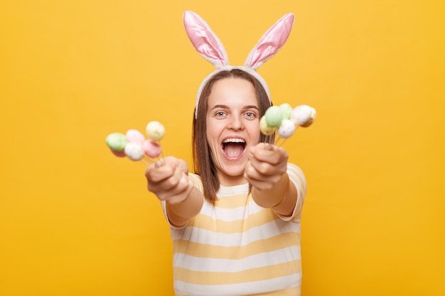 Imagem de uma mulher animada e festiva vestida de orelhas de coelho segurando mostrando bolo de Páscoa isolado sobre fundo amarelo gritando com cara positiva aproveitando o feriado