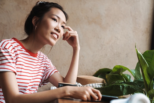 Imagem de uma mulher asiática bonita jovem alegre positiva sentar no café, olhando de lado.